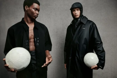 Two male models in black outerwear holding white soccer balls against a grey concrete wall background