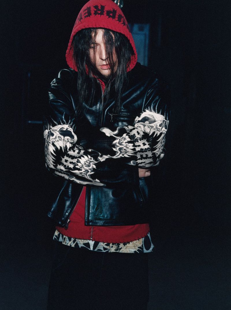 Person with long black hair wears a red hooded sweatshirt, leather jacket with arm designs, and skirt against a dark background.