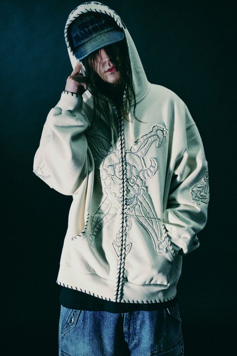 Person wearing a beige hoodie with embroidery and a denim cap posing in a dark studio setting with long hair partially covering their face.