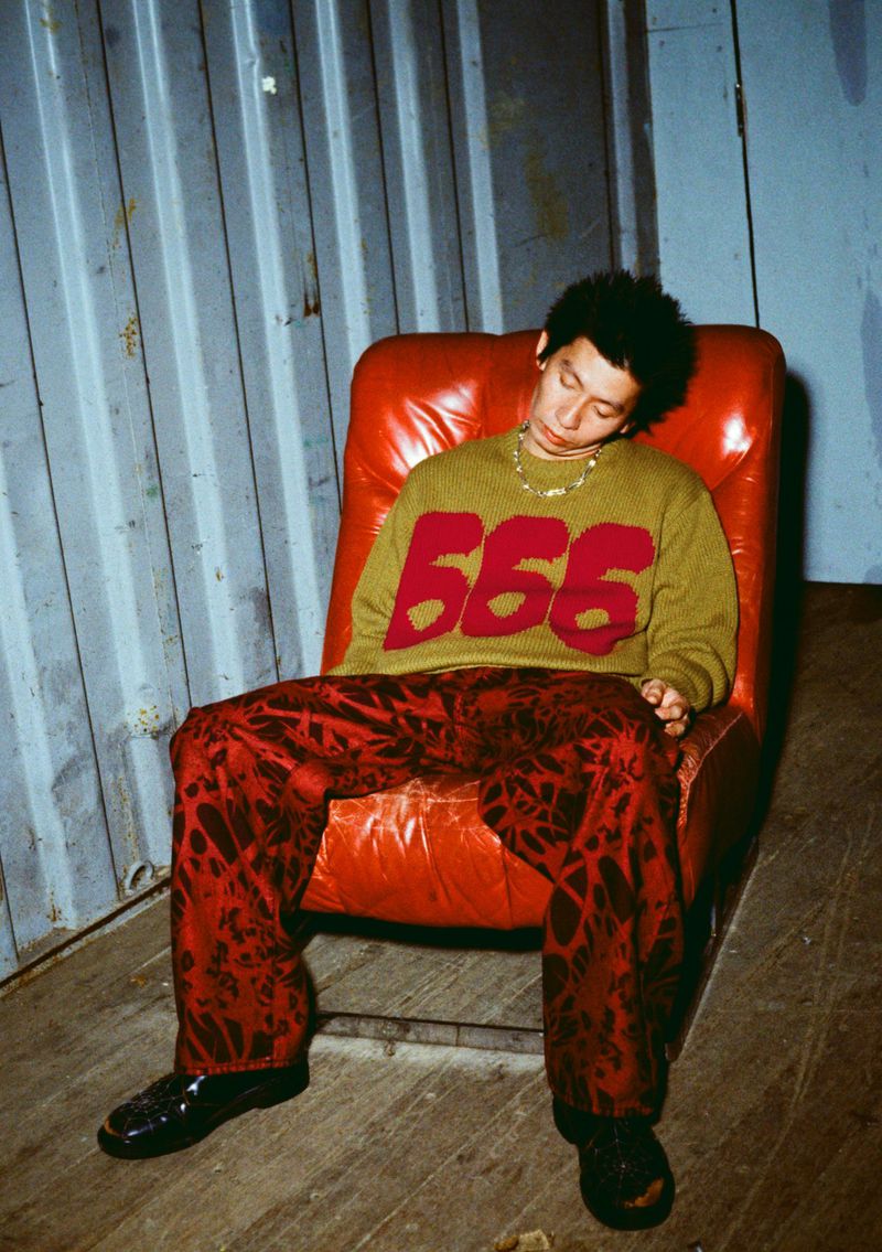 An adult male sits in an orange leather chair with his eyes closed, wearing patterned pants and a '666' sweater against a metal wall.