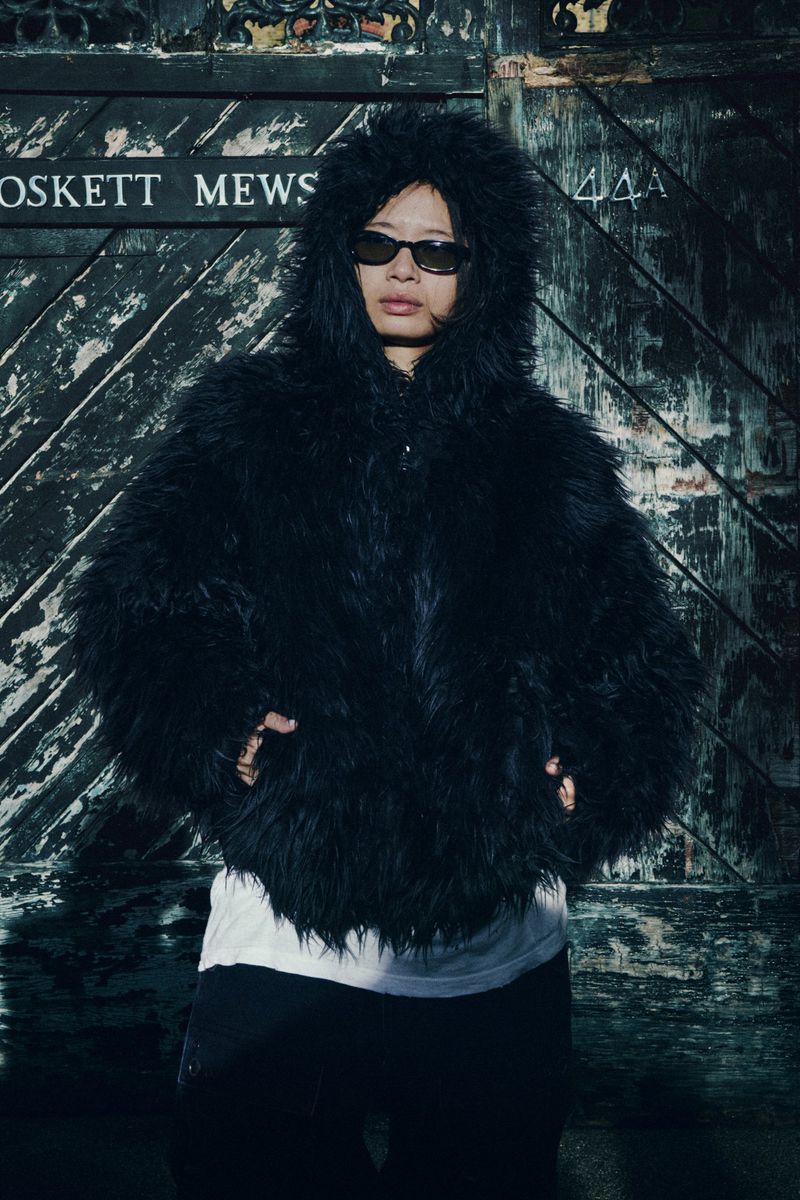 Person in oversized fur coat and sunglasses standing in front of a weathered door with signage, creating a unique fashion statement.