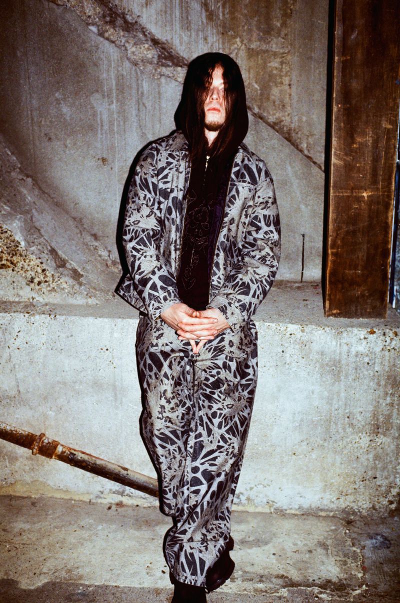 Full shot of a person in a grey and black patterned outfit with a scarf, posing in front of a concrete wall, looking at the camera.