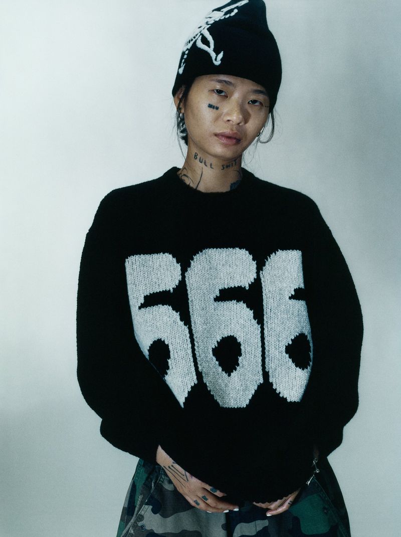 Stylish portrait of an adult female wearing a black knitted hat and sweater with the number 566, standing against a neutral background.