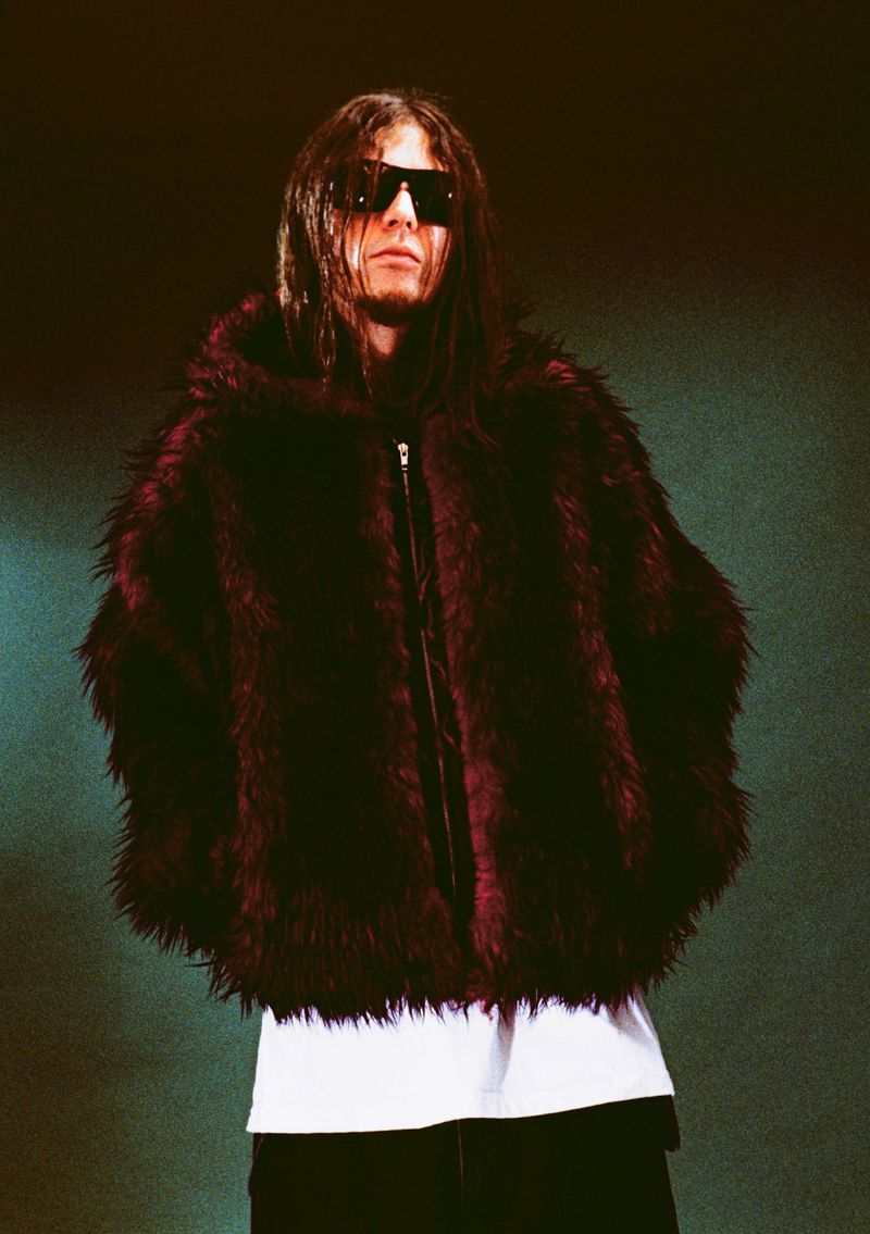 Person with long dark hair and sunglasses wearing a large, maroon faux-fur hooded coat in a medium shot against a dark background.