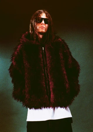 Person with long dark hair and sunglasses wearing a large, maroon faux-fur hooded coat in a medium shot against a dark background.