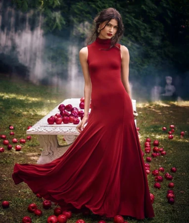 Elegant woman in a flowing red gown stands near a stone bench covered in red apples in an outdoor garden setting with soft lighting.