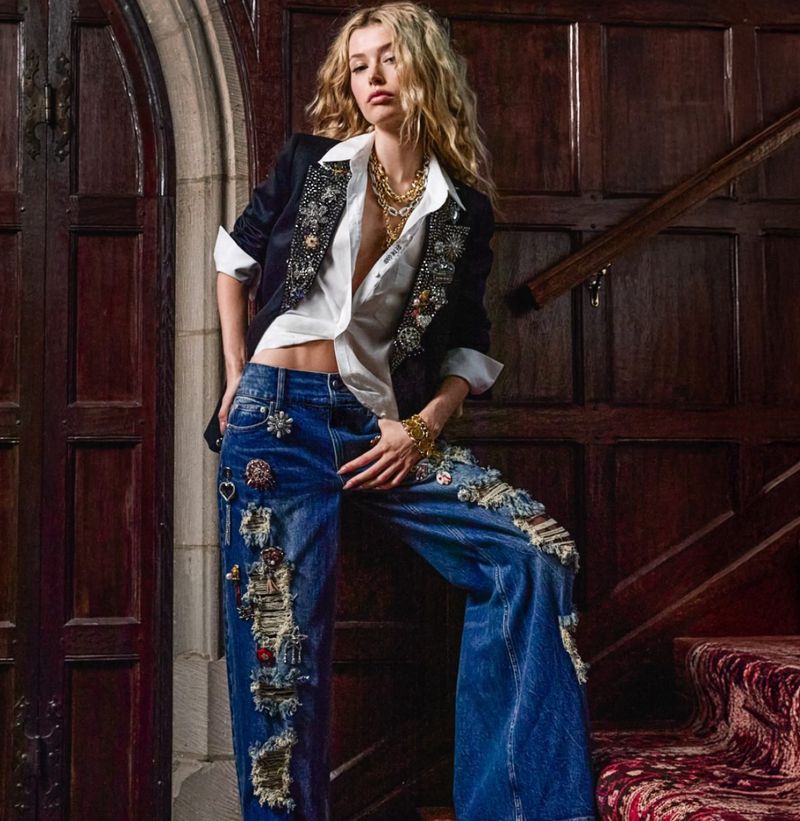 Blonde female model in ripped jeans and a bedazzled jacket poses indoors. She wears a gold necklace and bracelet, with a vintage door.