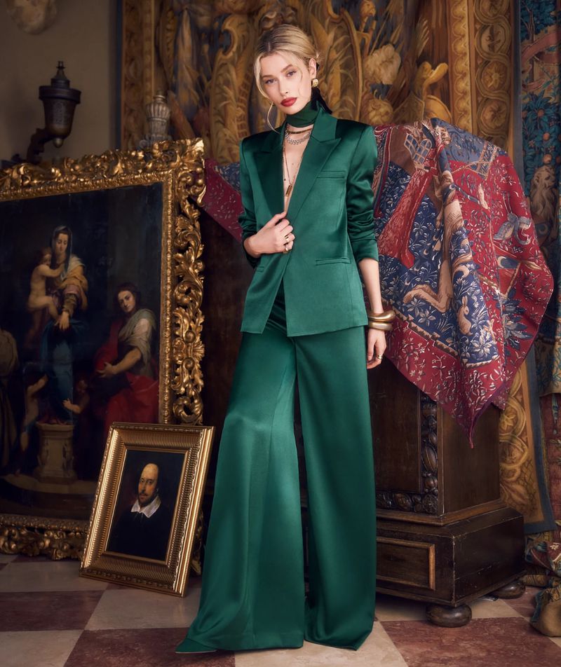 Elegant blonde woman in a green satin suit stands in an ornate interior with paintings and patterned textiles.