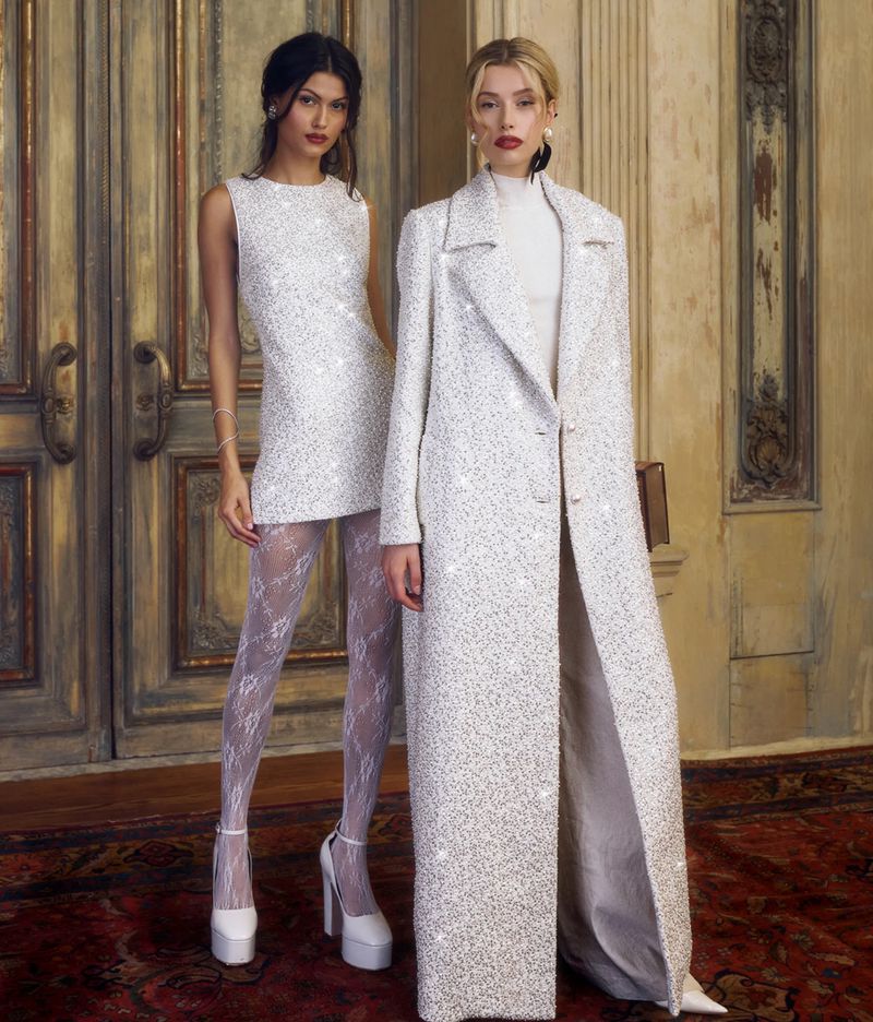 Two fashion models pose indoors wearing elegant white, textured dresses and coats with a vintage door as a backdrop.