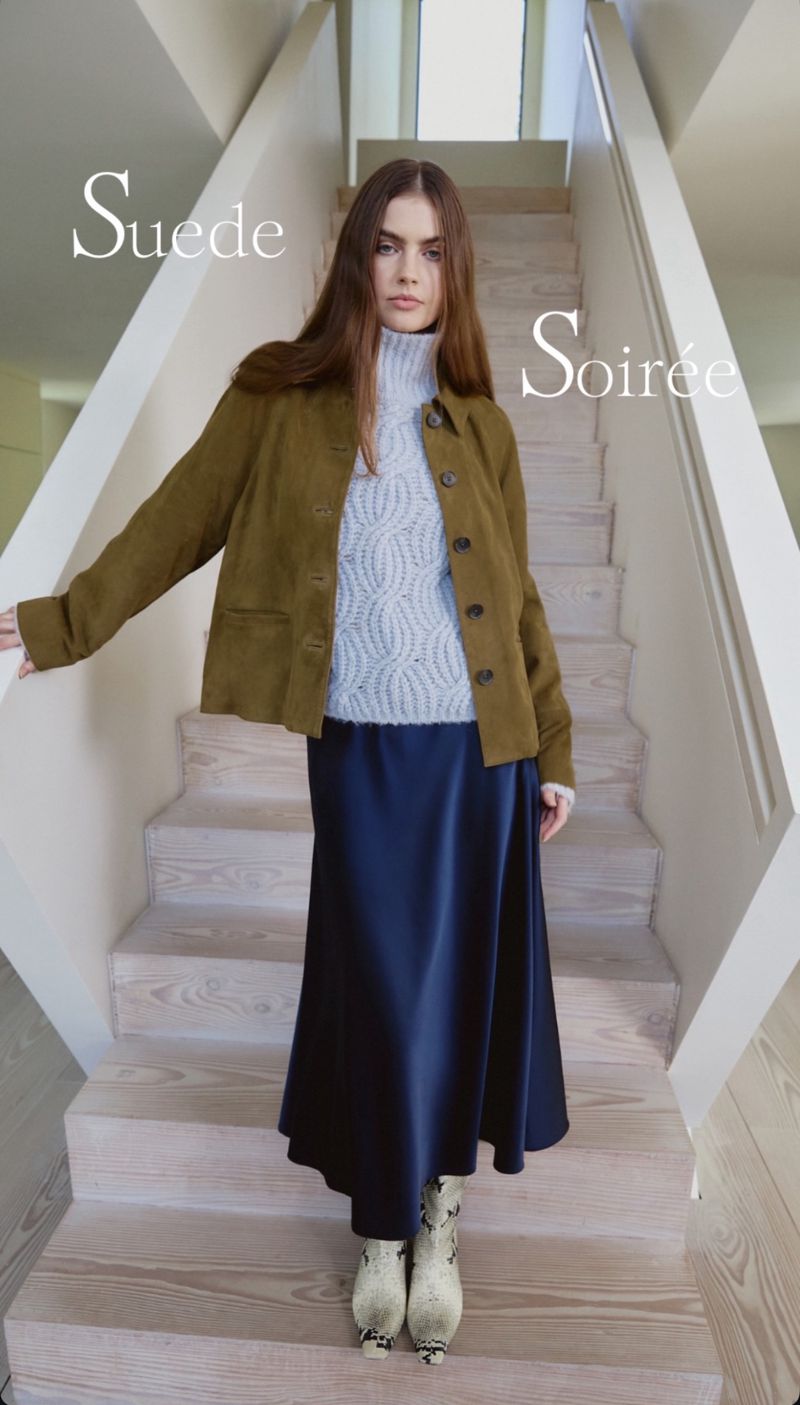 A brunette woman poses on stairs wearing an olive jacket, light-blue patterned sweater, navy skirt, and printed ankle boots inside.