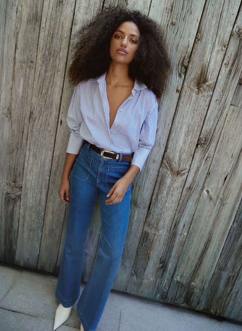 A slender woman with voluminous dark curly hair poses in jeans and a pinstripe shirt against a weathered wood wall for a fashion shot.