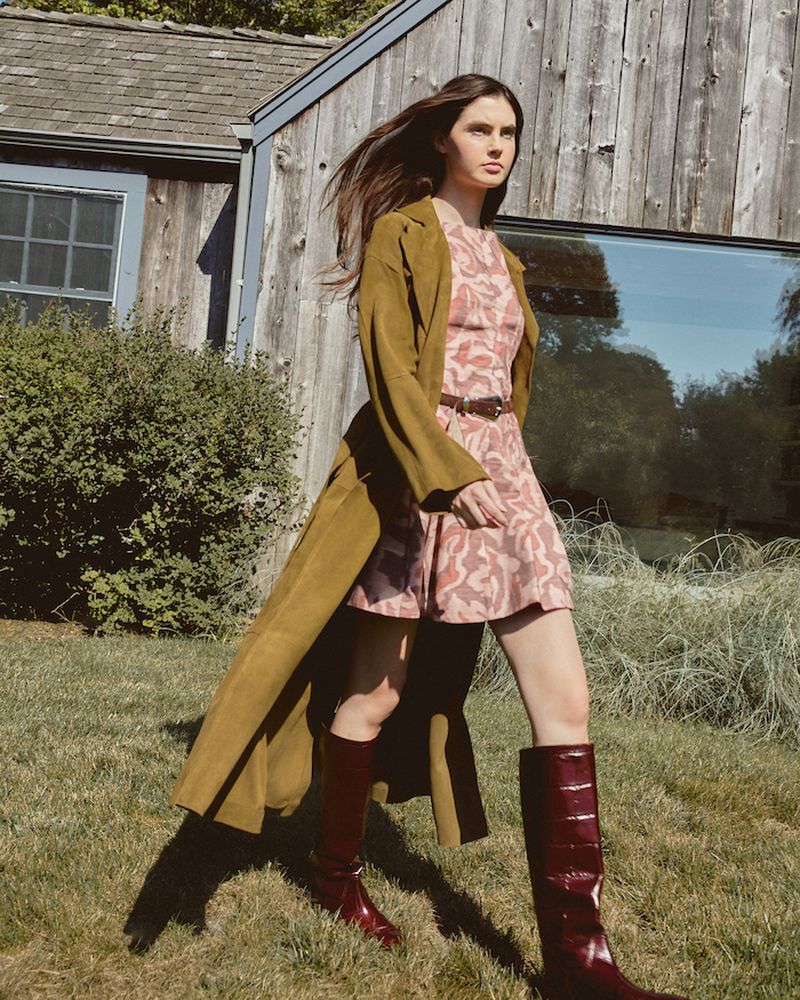 A brunette woman strides across a grassy lawn wearing a patterned mini dress, long coat, and boots, with a wooden building behind.