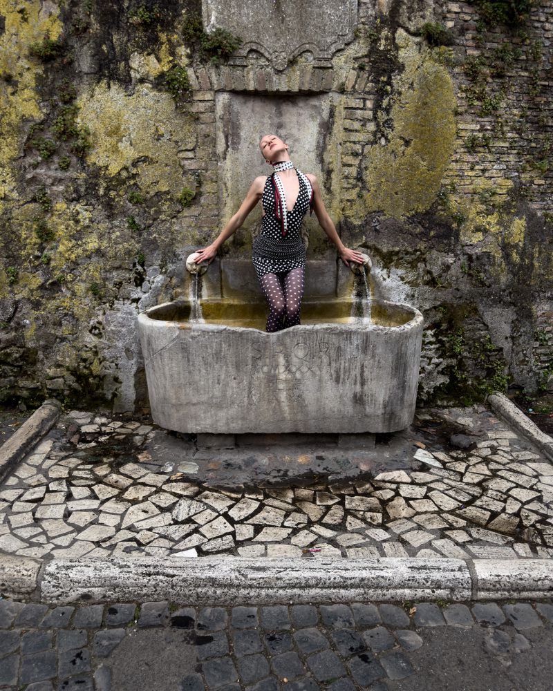 A woman stands waist-deep in a stone fountain with water, wearing polka dots, against a mossy brick backdrop outdoors.