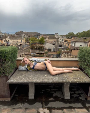 Model lying on a stone rooftop bench wearing patterned swimwear and a jacket against a cityscape view under an overcast sky.