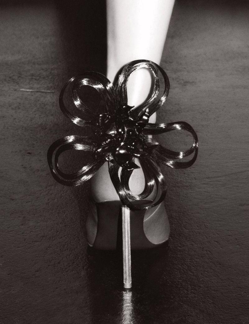 Close-up of an ankle-strap high-heel shoe with a sculptural flower embellishment on a reflective dark gray surface; studio shot.