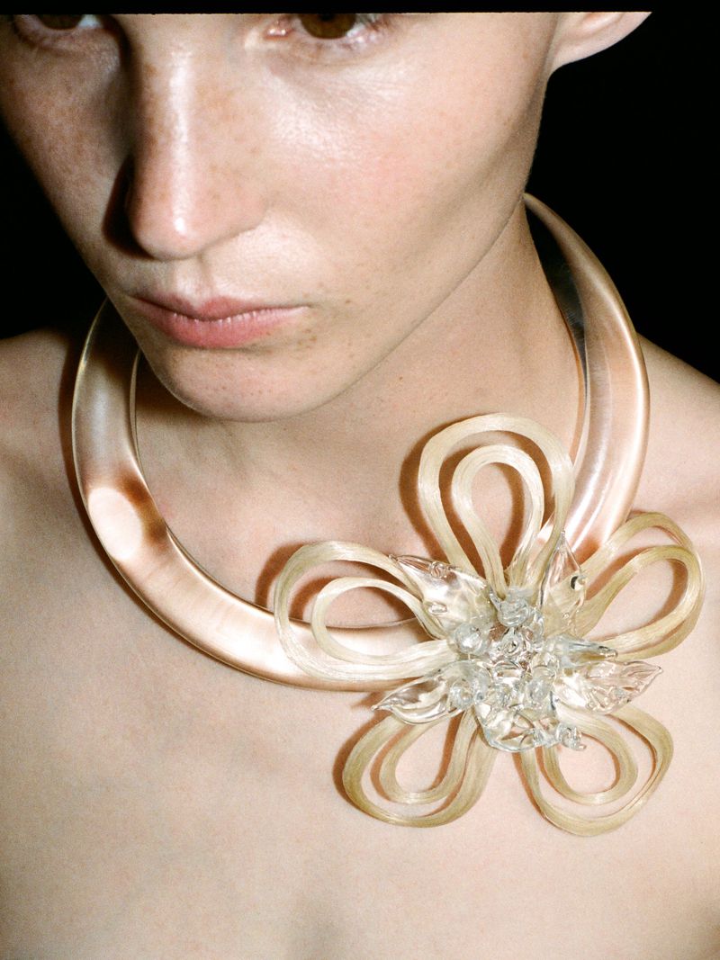 Close-up portrait of a fair-skinned adult female wearing a large, transparent, sculptural necklace with a flower-like design.