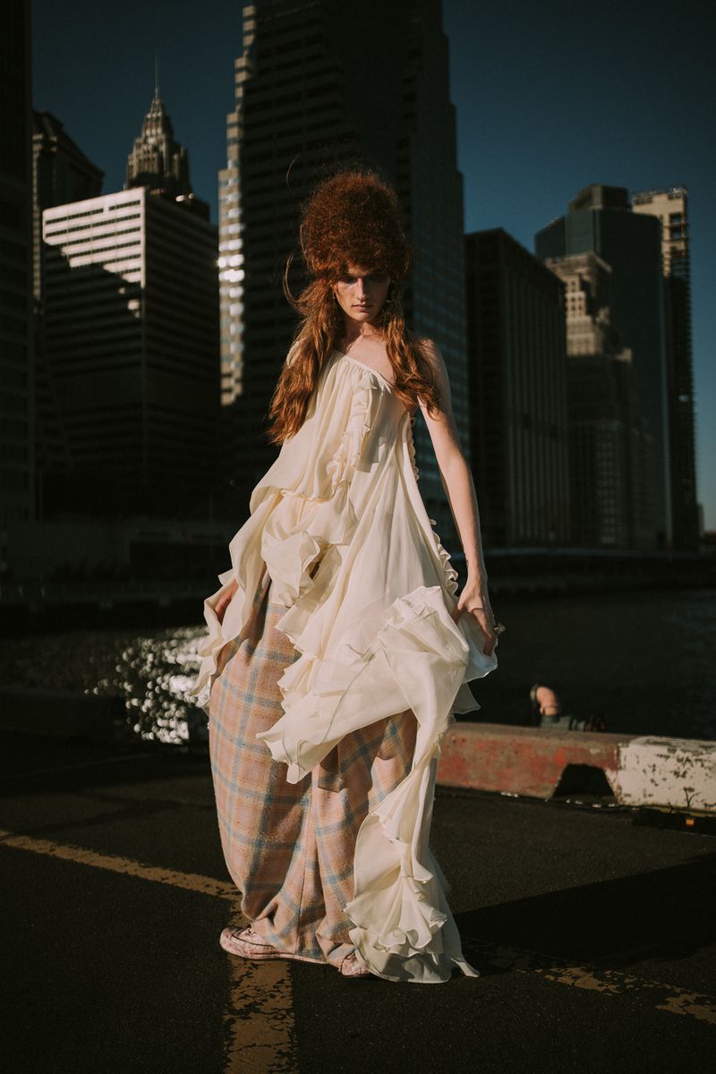 A slender female fashion model poses in a flowing white dress in an urban setting with high-rise buildings and a river backdrop.