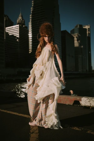 A slender female fashion model poses in a flowing white dress in an urban setting with high-rise buildings and a river backdrop.