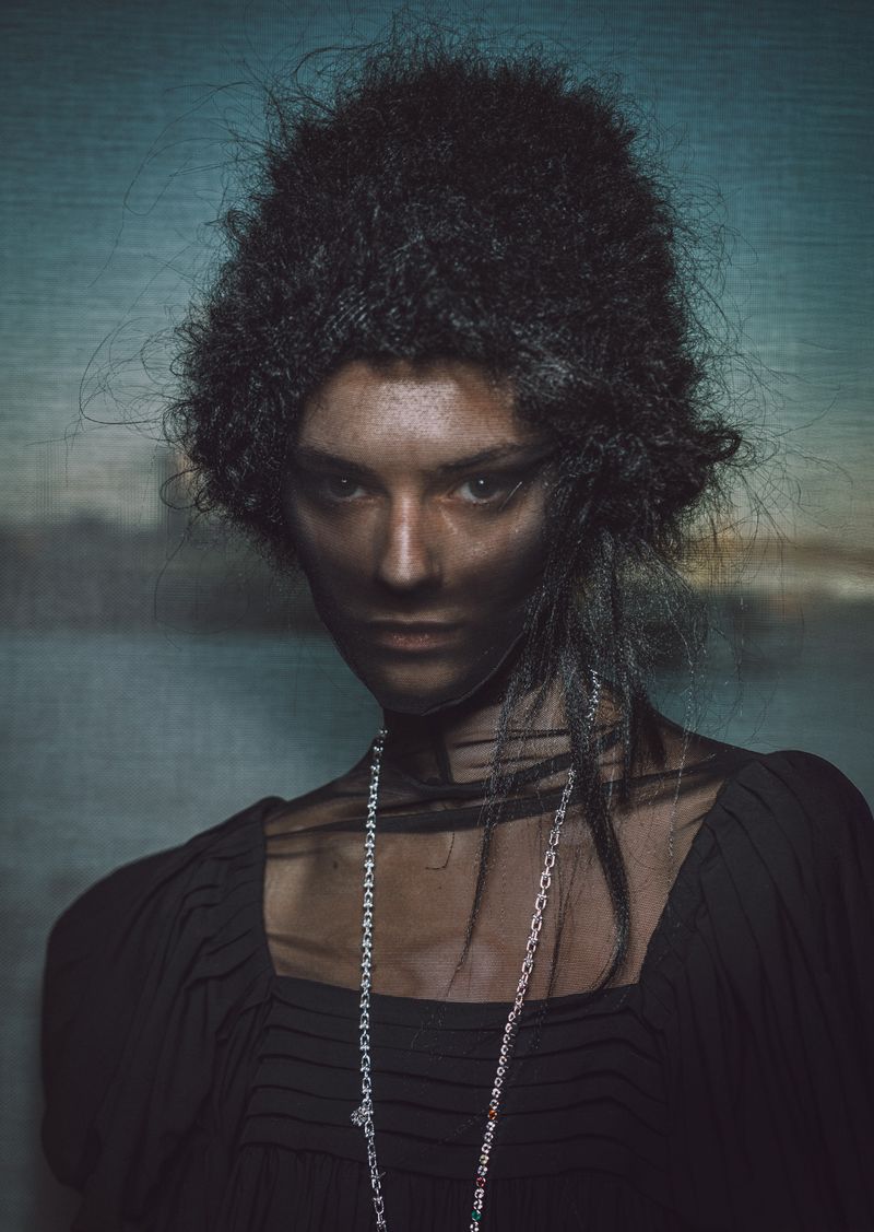 Fashionable woman with afro hairstyle poses in a dark dress and long silver necklace against a textured blue and white studio backdrop.