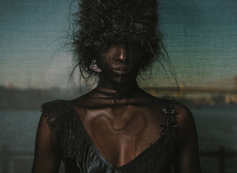 Dark-skinned woman with voluminous afro hairstyle, wearing a black lace top, choker, and an earring, poses against a muted backdrop.