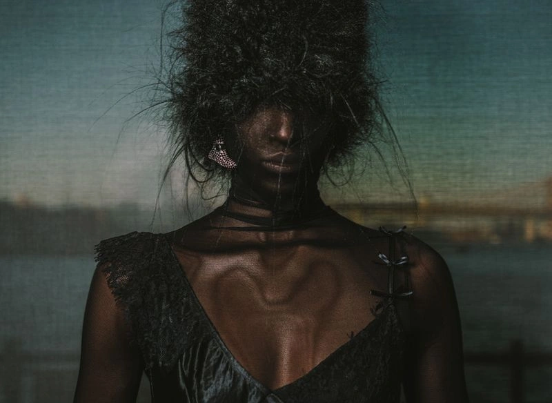 Portrait of a model with voluminous dark hair covering eyes, wearing a black garment and sparkling earring for Collina Strada.