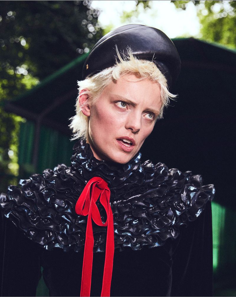Headshot of a short blonde-haired woman wearing a black hat and ruffled collar with a red ribbon, gazing upward in an outdoor setting.