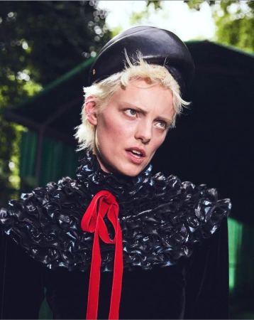 Headshot of a short blonde-haired woman wearing a black hat and ruffled collar with a red ribbon, gazing upward in an outdoor setting.