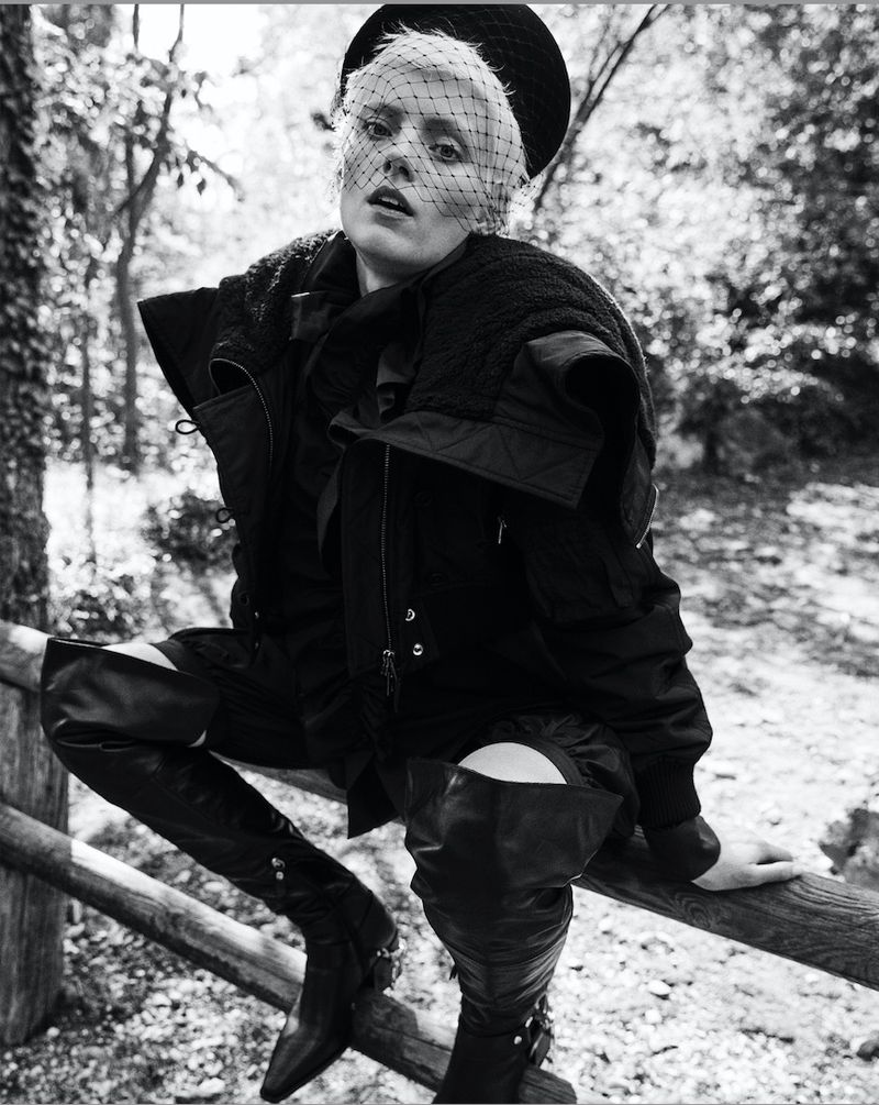 Black and white portrait of a person with short blonde hair, wearing a veiled hat and leather boots, sitting on a wooden rail outdoors.
