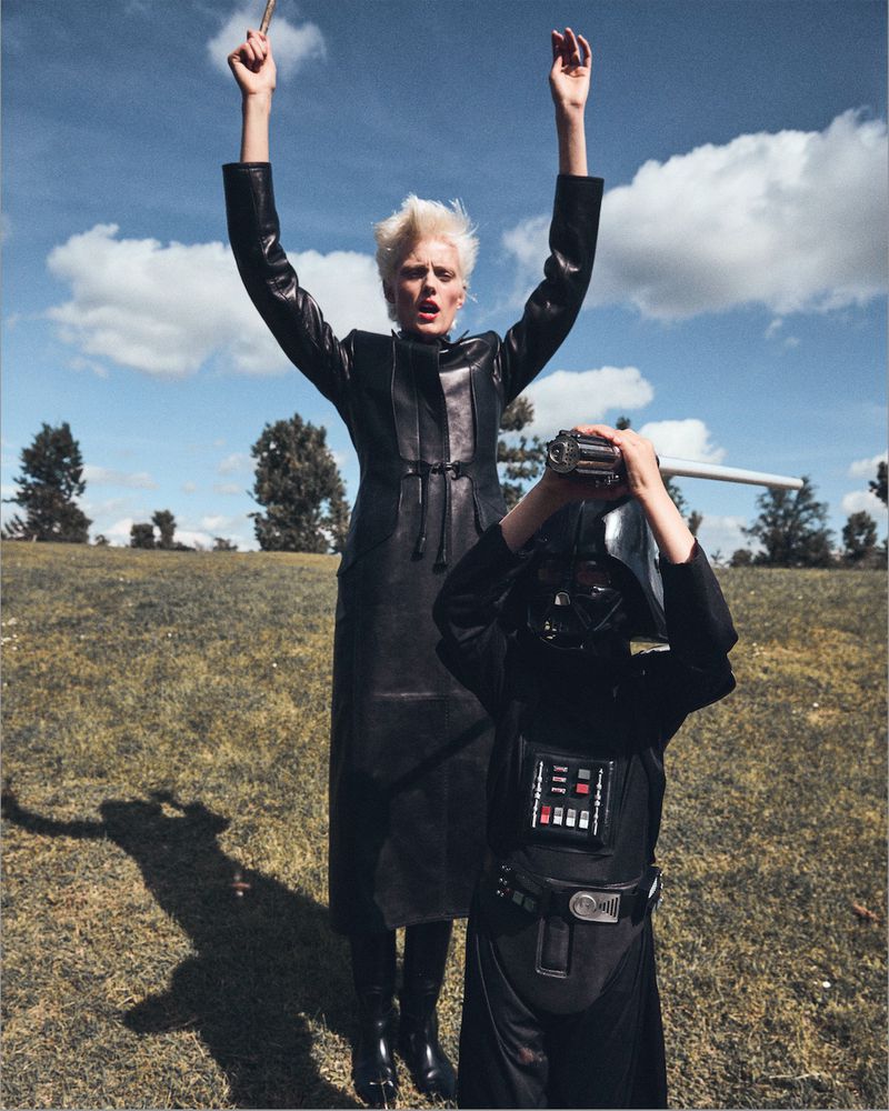 Adult female and child in Darth Vader costume with lightsaber in an outdoor field against a blue sky with white clouds.