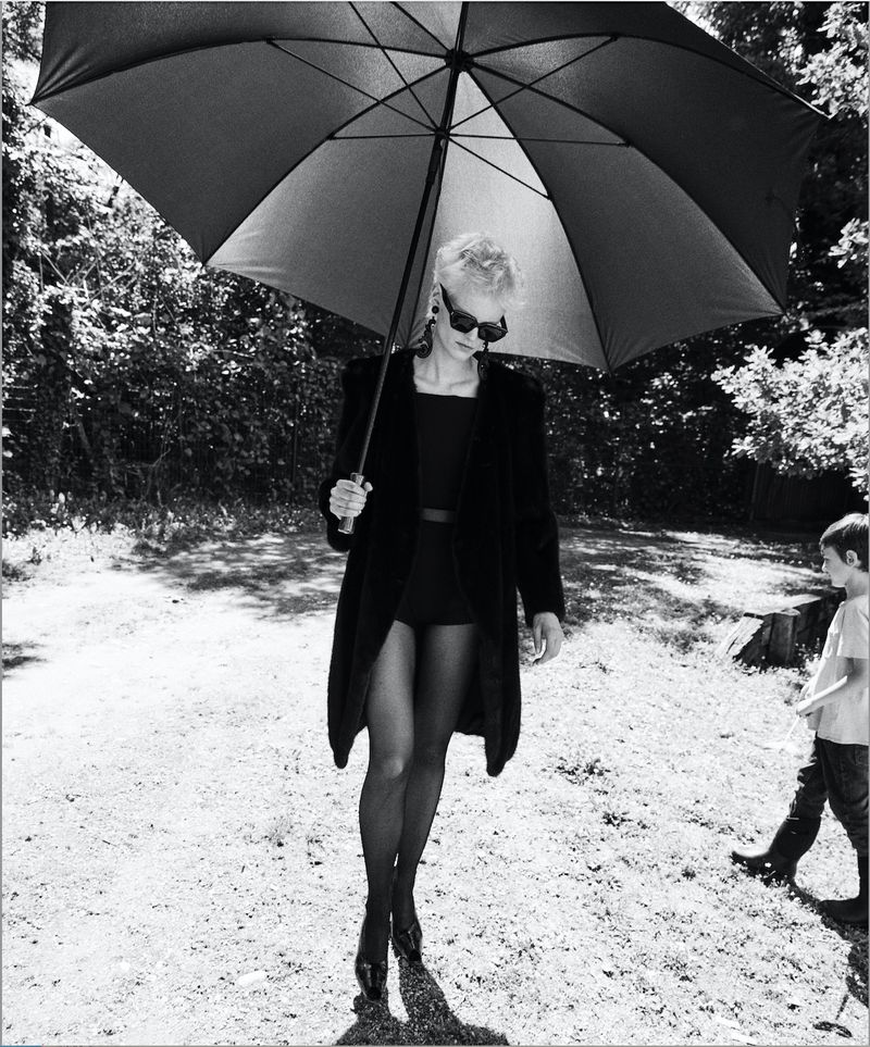 Woman in black coat and sunglasses holds umbrella on path, boy in background. Black and white fashion shot in outdoor setting.