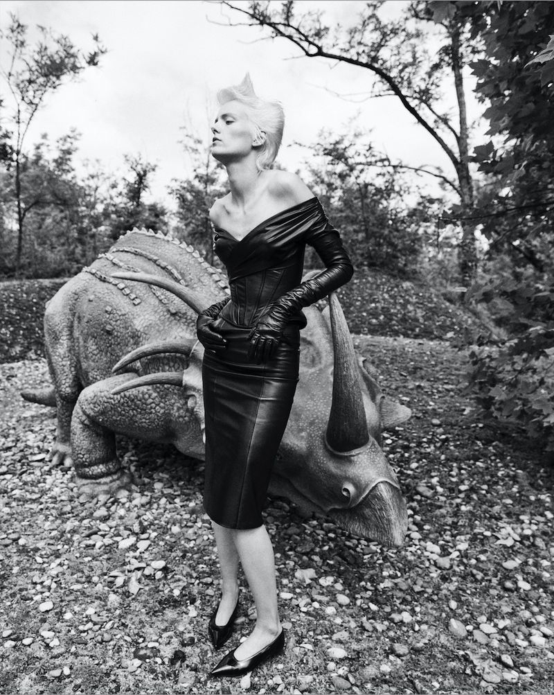 A black and white fashion shot of a woman in a black leather outfit posing next to a large Triceratops statue in an outdoor setting.