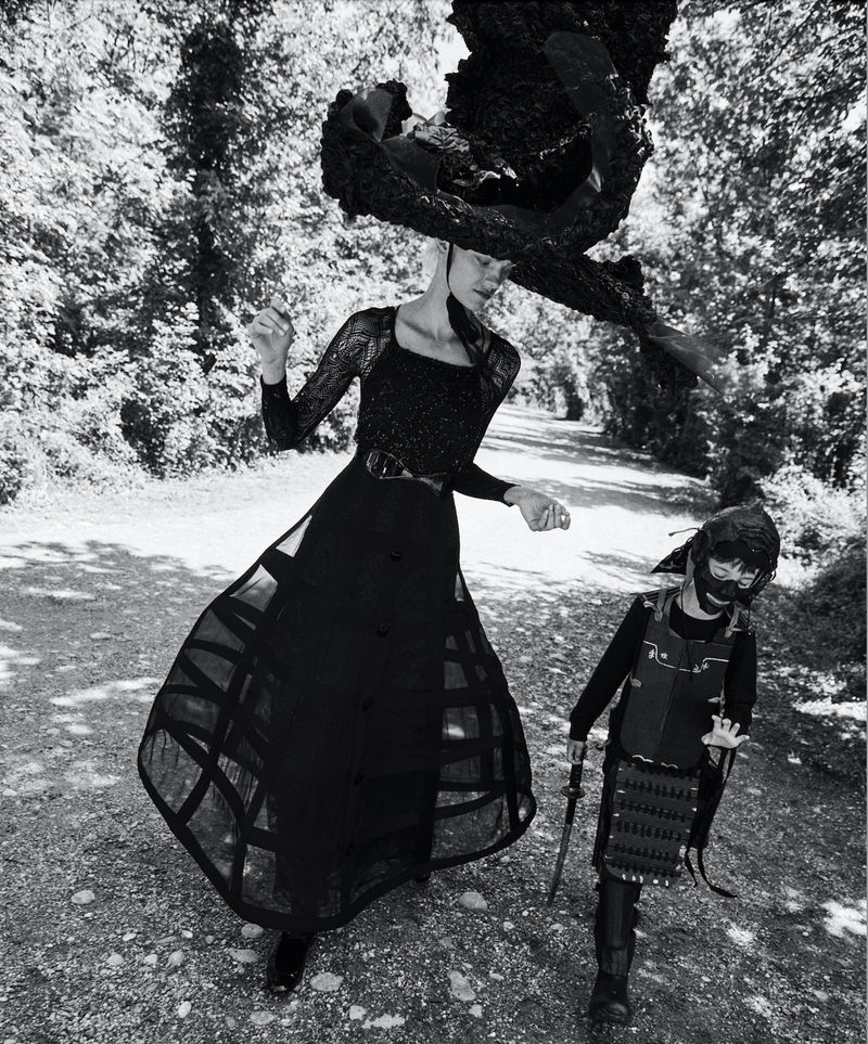 Black-and-white editorial fashion shot of an adult woman in long dress and large hat and a child in samurai costume in wooded landscape.