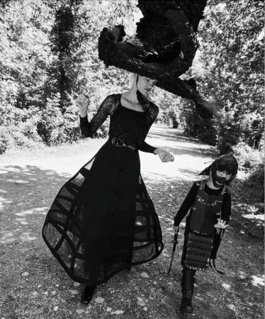Black-and-white editorial fashion shot of an adult woman in long dress and large hat and a child in samurai costume in wooded landscape.
