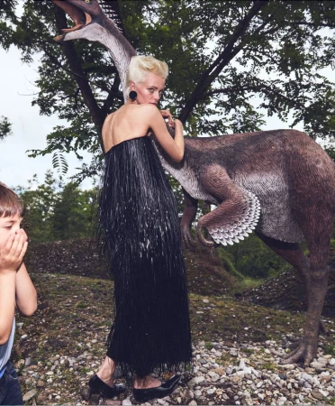 A woman in a black dress poses with a dinosaur statue in an outdoor setting, while a young child covers their face with their hands.
