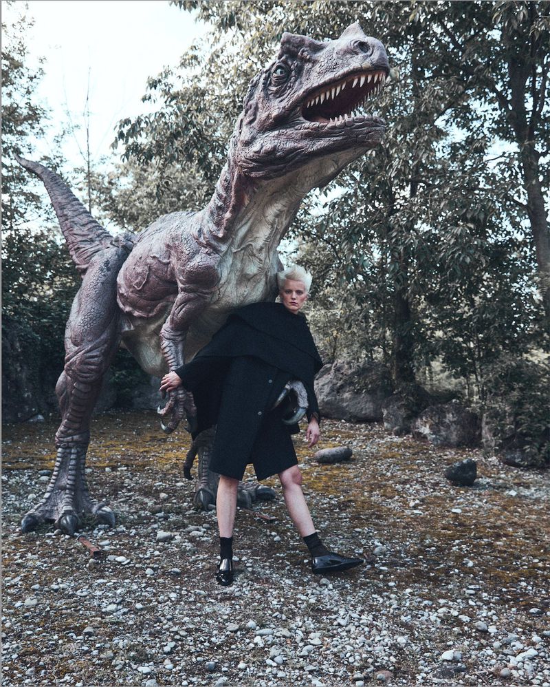 Woman in black outfit poses next to a large dinosaur statue in a forest setting with sparse foliage and a ground covered in pebbles.