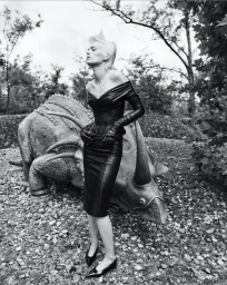 Model Maaike Klaasen in a black leather dress posing beside a triceratops sculpture outdoors in a wooded setting.