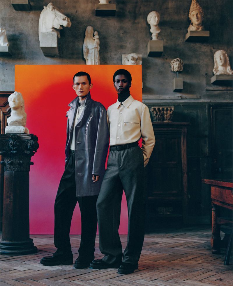 Two male fashion models pose in front of a red-orange gradient backdrop, surrounded by classical sculpture in a vintage architectural space.