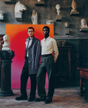 Two male fashion models pose in front of a red-orange gradient backdrop, surrounded by classical sculpture in a vintage architectural space.