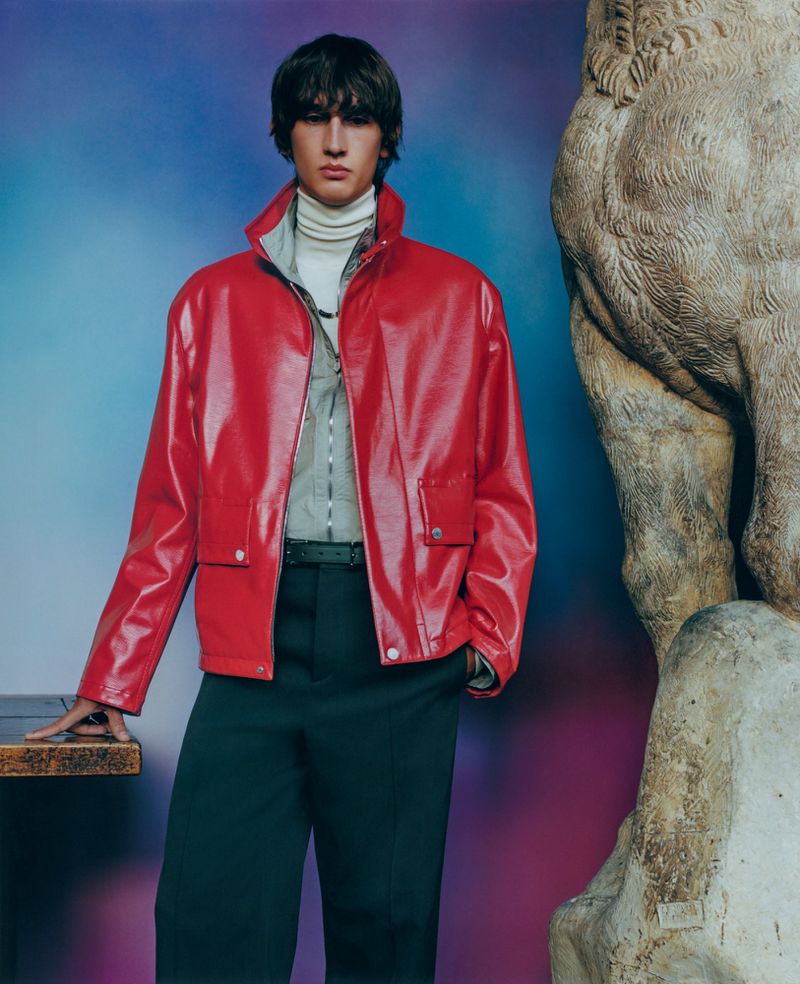 Adult male poses indoors wearing a red jacket, dark pants, and white turtleneck, standing next to a carved stone sculpture.
