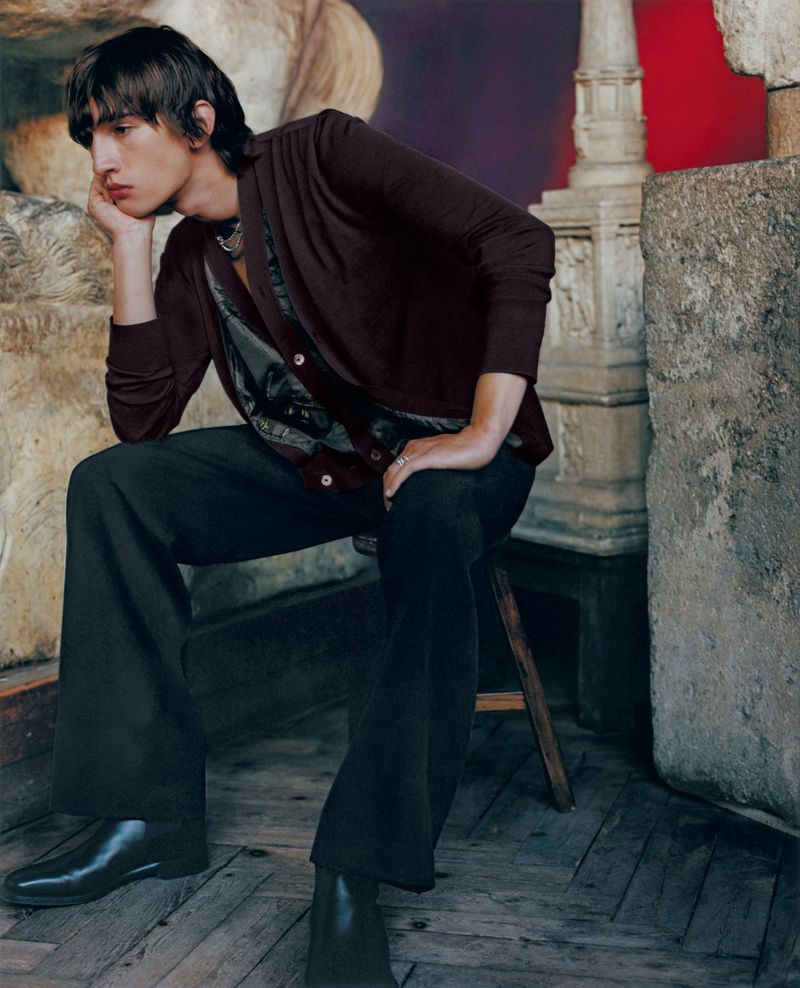 A young adult male poses indoors wearing a burgundy cardigan, dark pants, and black boots while seated on a wooden stool in a studio.