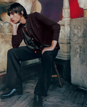A young adult male poses indoors wearing a burgundy cardigan, dark pants, and black boots while seated on a wooden stool in a studio.