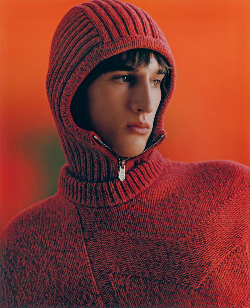 Close-up studio shot of a young adult male with brown hair wearing a zip-up hooded red knit sweater against an orange gradient background.