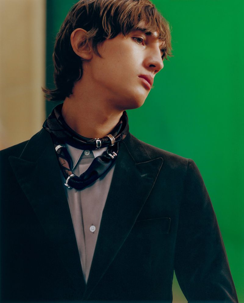 Young man in a dark blazer and decorative tie stands against a vivid green background in a medium studio shot. Fashion photography.