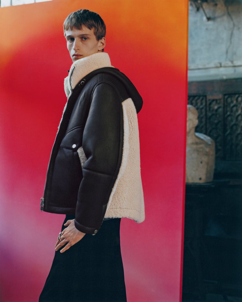 Adult male fashion model poses in a hooded brown and white jacket against a red gradient backdrop in an indoor environment.