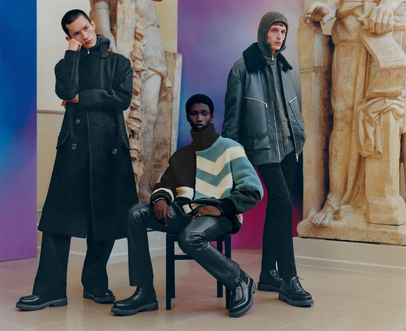 Three adult male fashion models pose indoors wearing wool coats, jackets, sweaters, and leather pants in front of marble statue art.