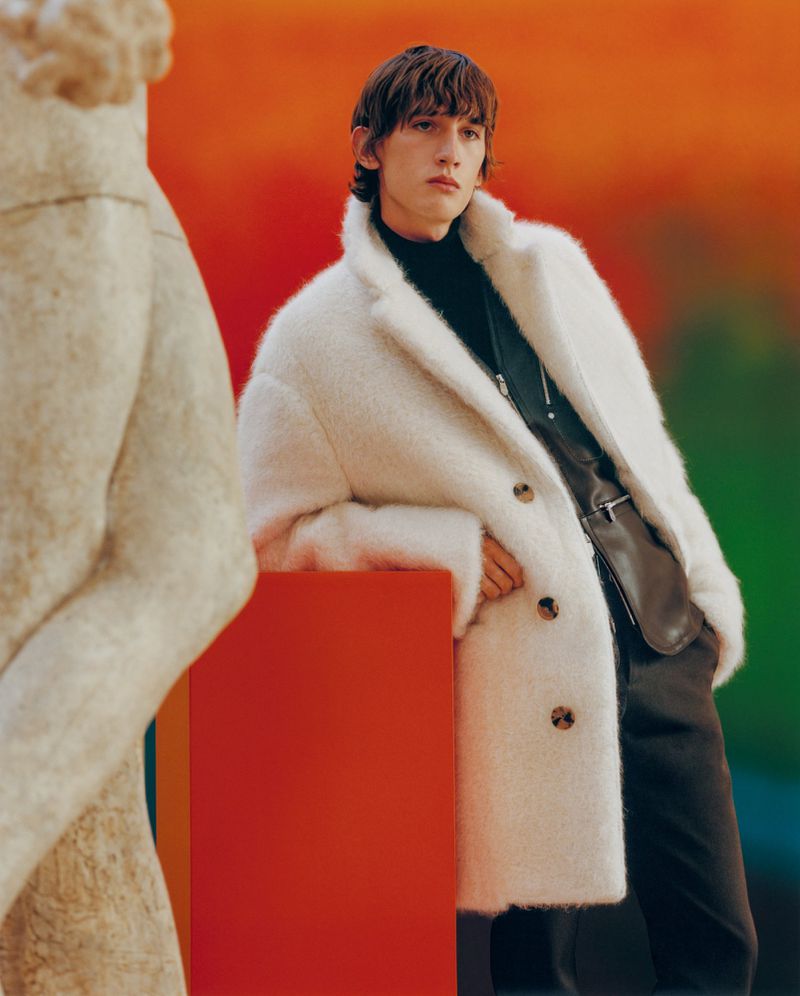 A male model poses in a fuzzy white coat, black turtleneck, and brown pants with a colorful background, next to a marble statue.