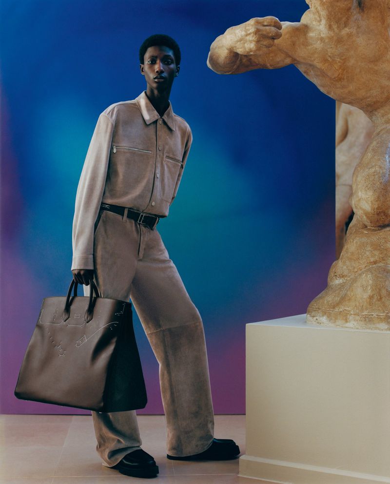 An African American male model poses with a handbag in a studio with bright blues and purples, beside a sculpture.