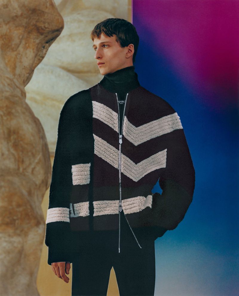 Fashion shot of a young adult male wearing a black turtleneck and black and white pattern jacket standing next to a stone statue.