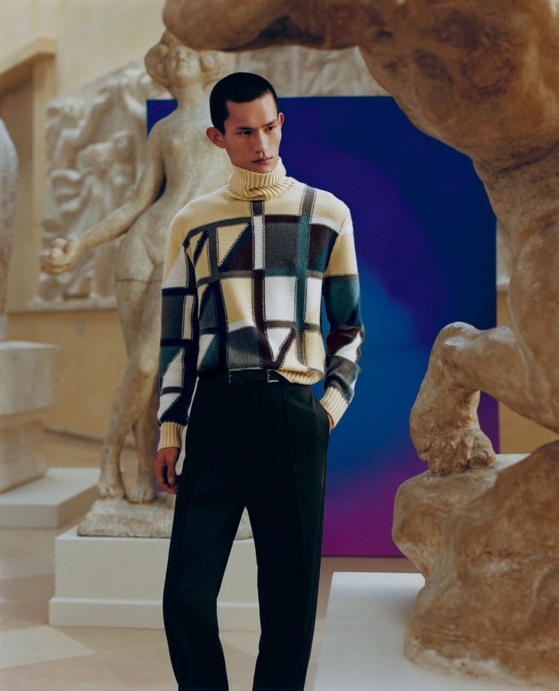 Male model wears patterned turtleneck sweater and dark pants among classical sculptures and a gradient background panel in an indoor setting.