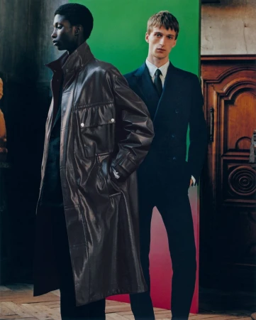 Two men pose in an indoor setting; one wears a brown leather coat, the other a suit, creating a stylish editorial fashion image.